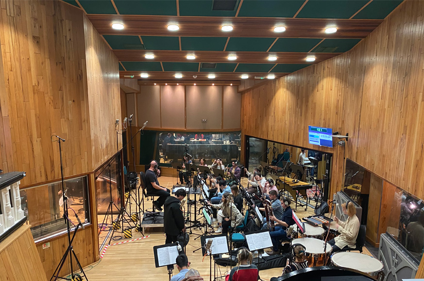 RCM Symphony Orchestra performing at Abbey Road Institute in a wood-panelled recording hall, with recording studio behind them.